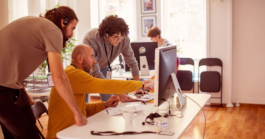 A group of three people collaborates around a desk with a computer, discussing a project. They appear focused and engaged in a friendly and productive environment.