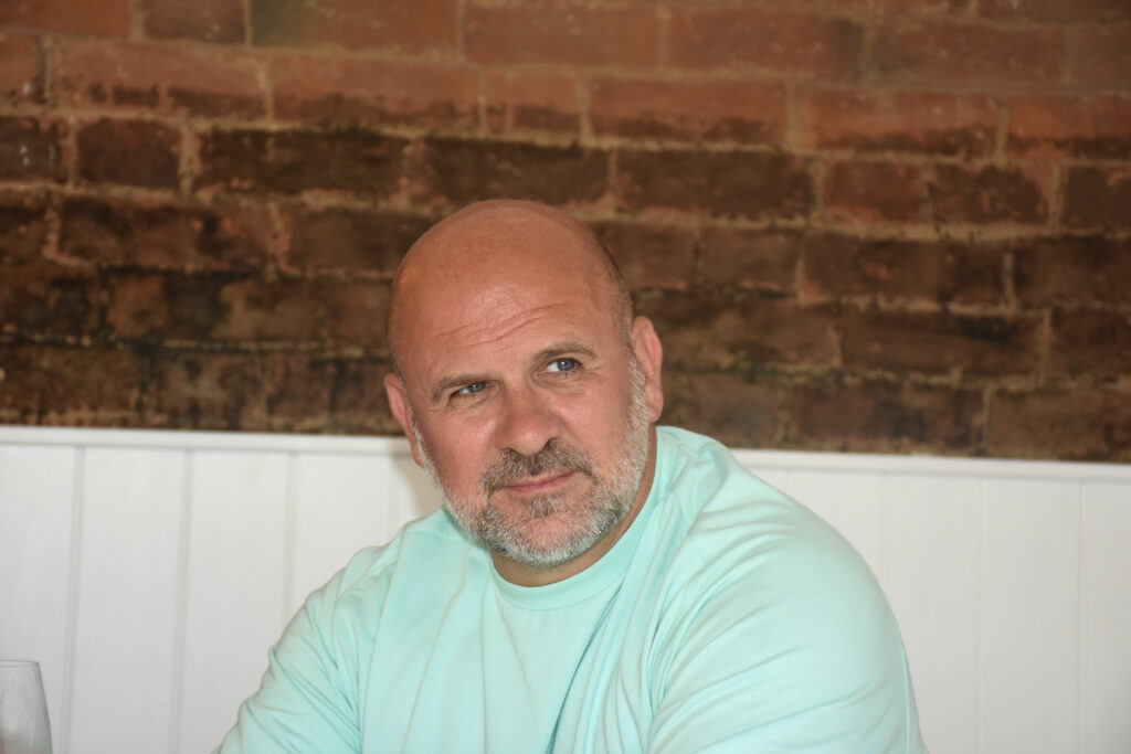 A man with a beard wearing a light blue shirt sits in front of a brick wall, looking off to the side.