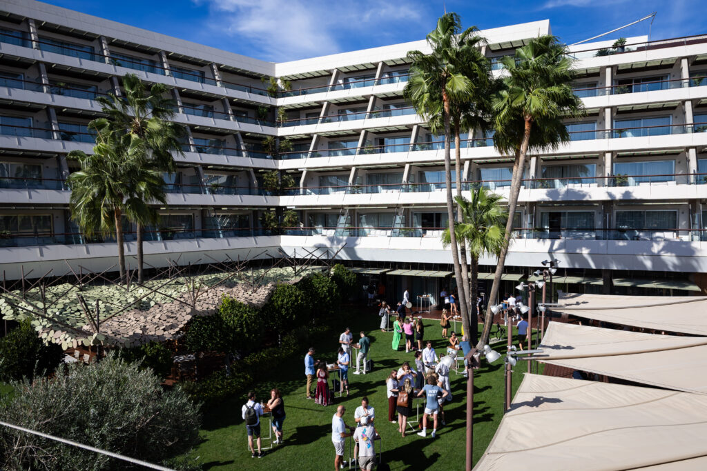 A modern hotel courtyard features lush palm trees and groups of people socializing on a green lawn, surrounded by a contemporary building with large windows under a clear blue sky.