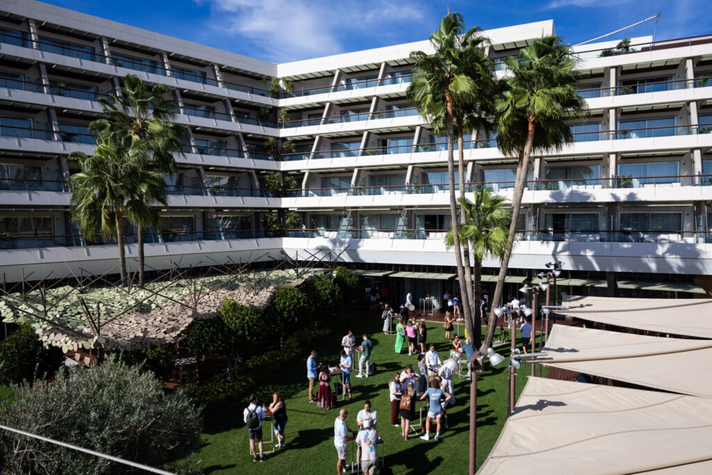 A group of people mingling on a sunny, grassy courtyard surrounded by a large, modern hotel building with several floors and palm trees.