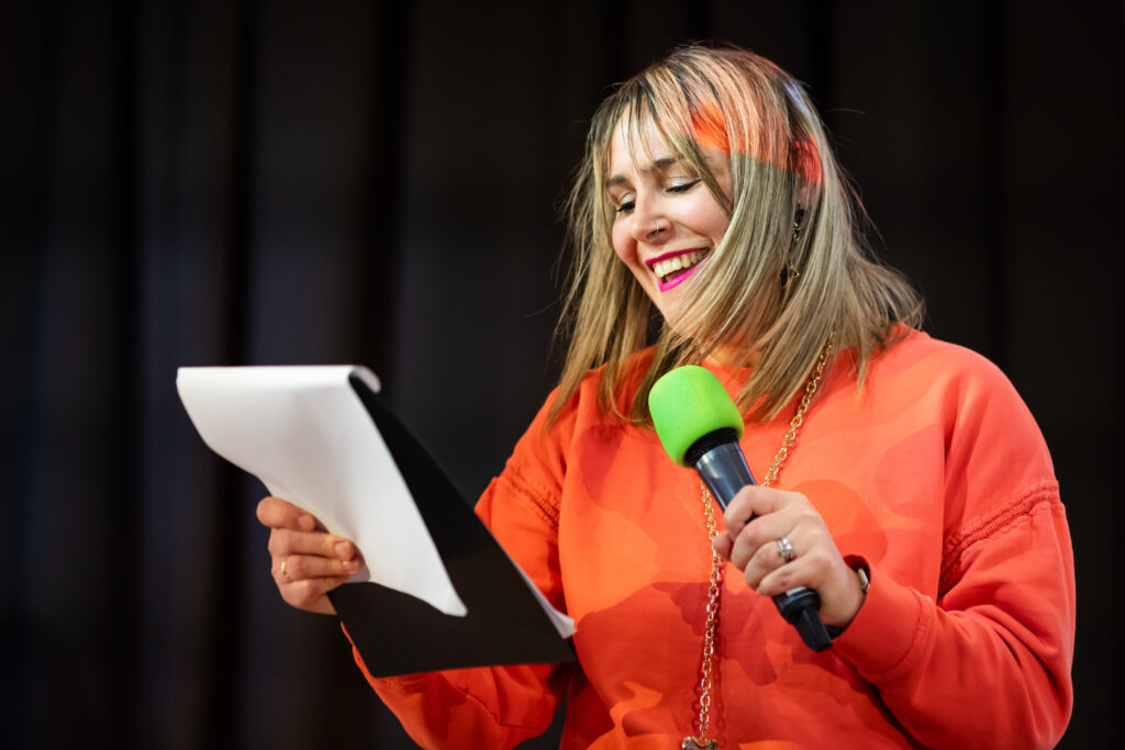 A smiling woman holds a clipboard with notes in one hand and a green microphone in the other. She is wearing an orange sweater and standing in front of a black curtain, engaging with an audience or reading from her notes.