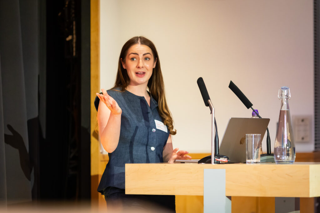 A woman with long brown hair presents at a podium, gesturing with her right hand. She is speaking to an audience, with a laptop and water bottle on the table. The setting appears to be a conference or seminar.