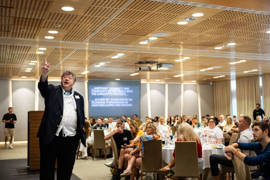 A speaker gestures passionately during a presentation in a conference room filled with seated attendees.