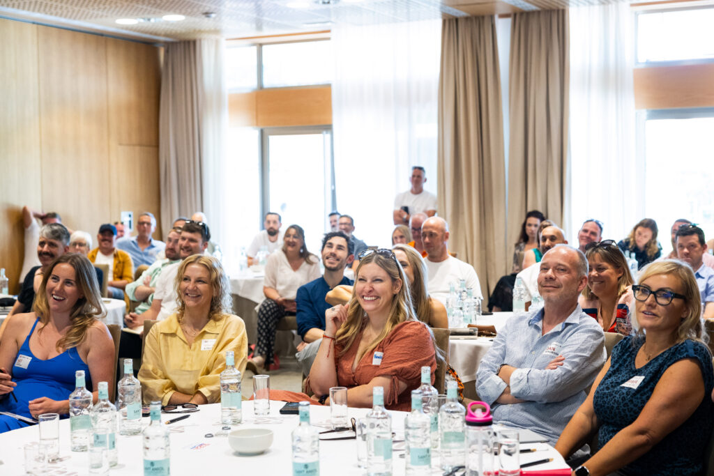 A group of people attentively listening to a presentation in a conference room, seated around tables with water bottles and notepads.
