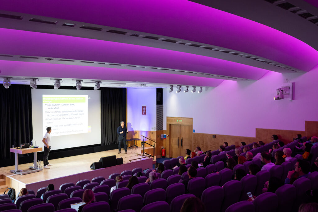 A speaker presents to an audience in a modern lecture hall with purple lighting. A projector displays information on a screen, while attendees sit in purple chairs, some taking notes or using devices.