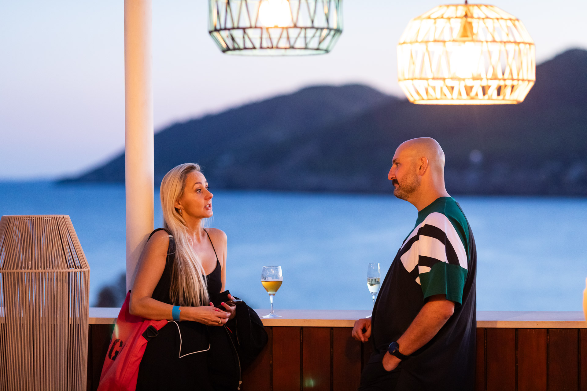 A woman with long blonde hair and a man with a shaved head converse beside a bar, overlooking a scenic ocean view at sunset. They each hold drinks, and decorative hanging lights add a warm ambiance to the setting.