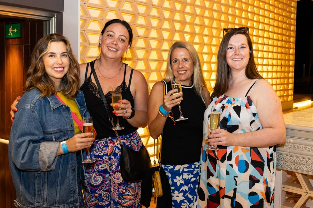 Four smiling women holding drinks, standing together in a warmly lit space with a geometric wall backdrop. They're dressed casually, suggesting a relaxed social gathering.
