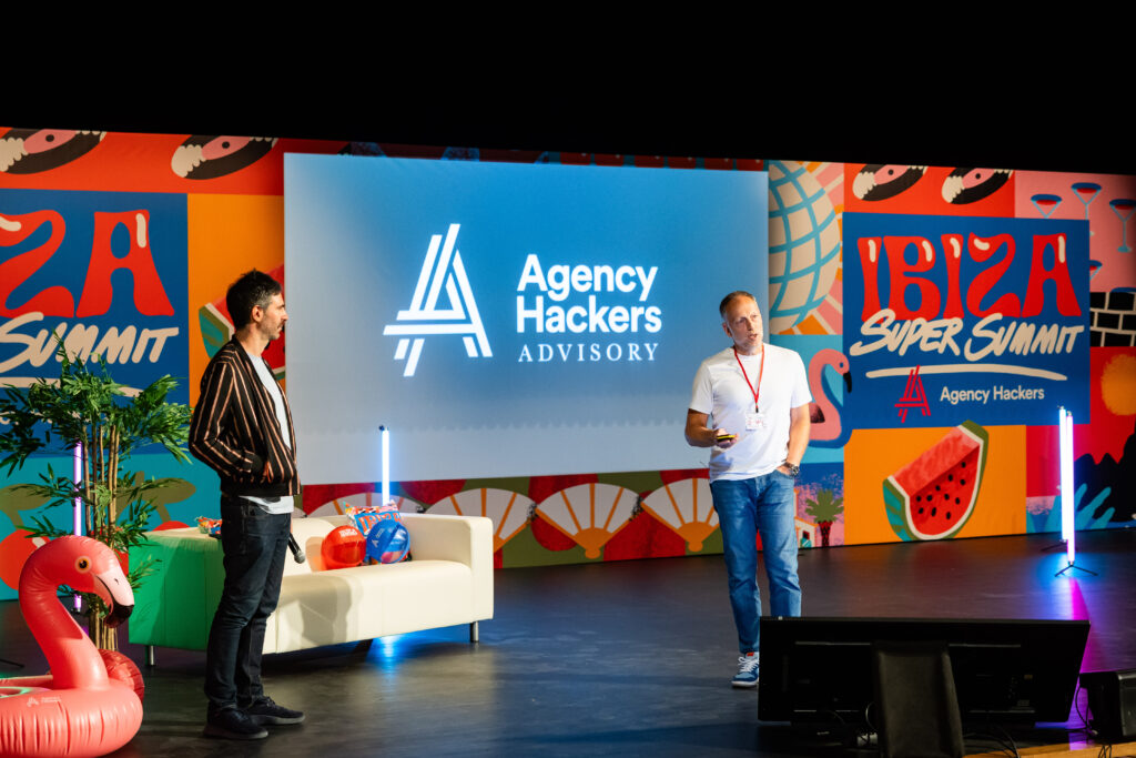 Two speakers present on stage at the Ibiza Super Summit, featuring a colorful backdrop with the Agency Hackers logo. The setting includes tropical decor and a casual atmosphere, highlighting an engaging discussion or presentation.