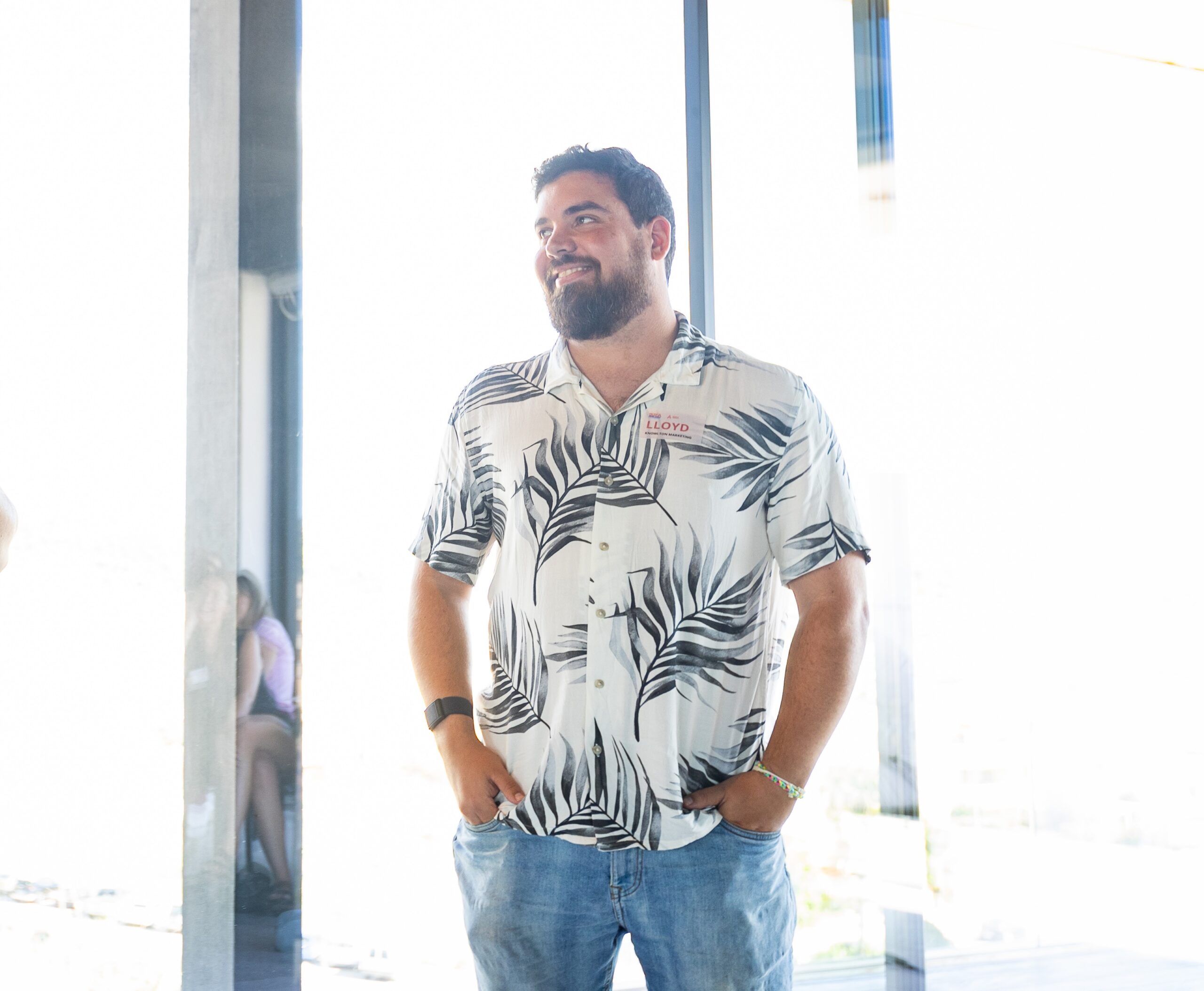 Man in a tropical leaf-patterned shirt and jeans stands near a window, looking to the side with a thoughtful expression.