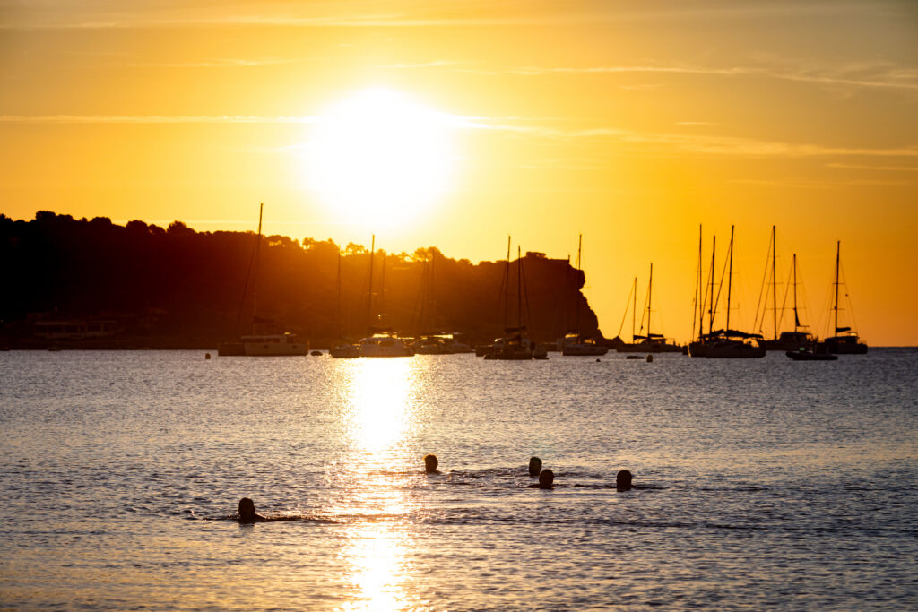 People swimming in calm waters during a vibrant sunset, with silhouetted boats and a rocky shoreline in the background.