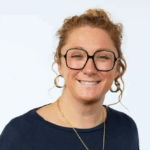 Person with curly hair, wearing large glasses and hoop earrings, smiling in a blue shirt against a plain background.
