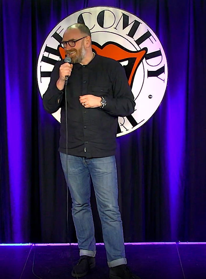 A comedian performs on stage, holding a microphone, with a backdrop featuring a comedy club logo.