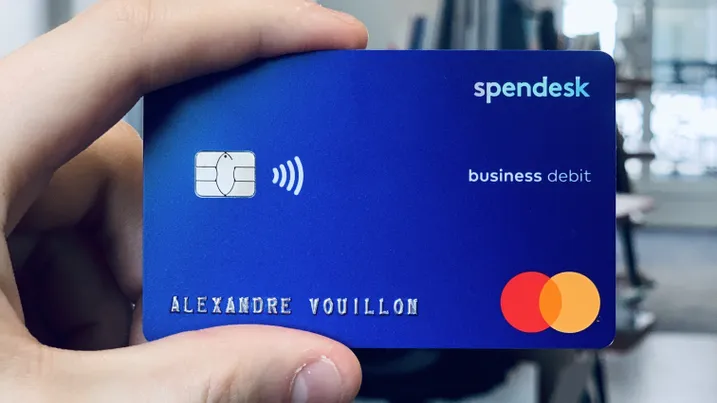 A hand holds a blue Spendesk business debit card featuring a chip and contactless symbol, with the Mastercard logo.