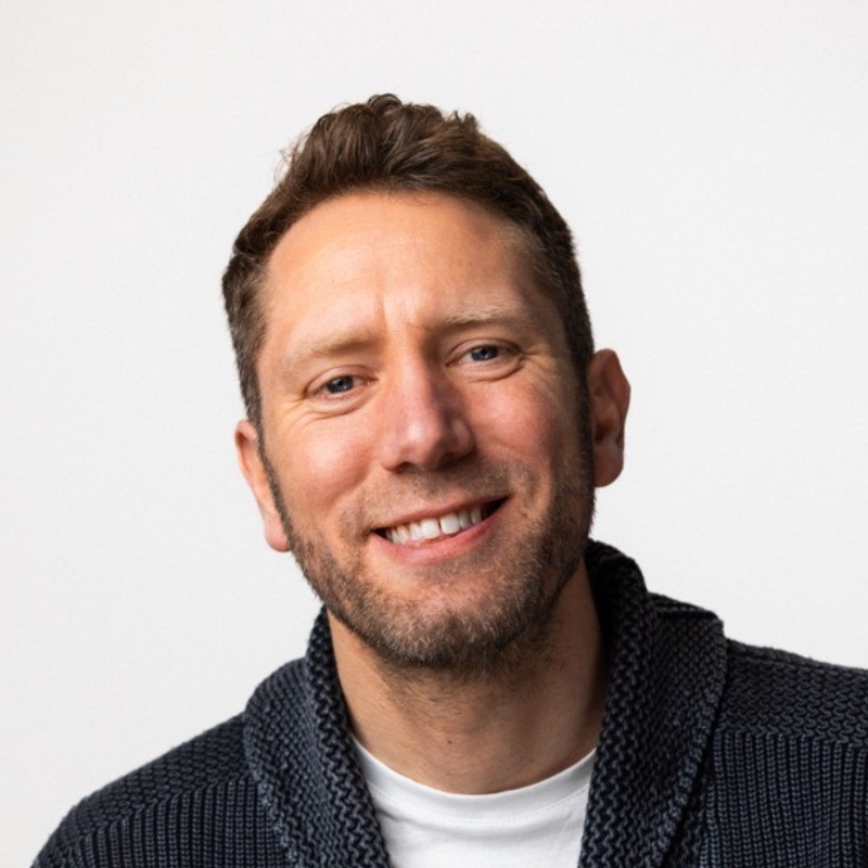Smiling man with short hair and a beard, wearing a dark sweater over a white shirt, against a plain white background.