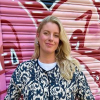 A person with long blonde hair is standing in front of a pink and red graffiti-covered wall, wearing a patterned jacket over a white shirt and smiling slightly.