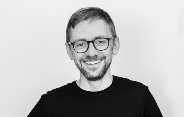 A smiling man with short hair and glasses wearing a black T-shirt against a plain white background.