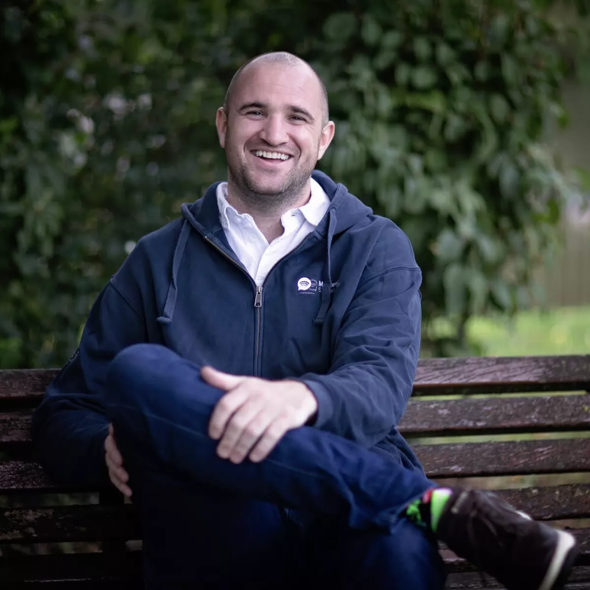 A man sitting on a bench outdoors, smiling, wearing a dark hoodie and jeans, with trees in the background.
