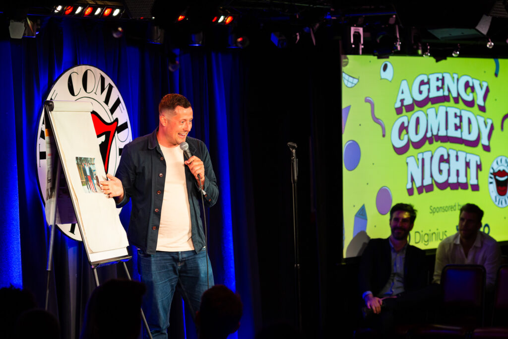 A comedian performs on stage holding a microphone and using a flip chart, with a sign saying "Agency Comedy Night" in the background and an audience watching.