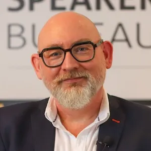 Bald man with glasses and a beard, wearing a white shirt and suit jacket, smiling in front of a blurred background with the word "BUREAU" visible.