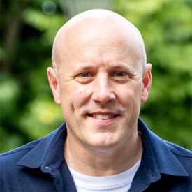 A bald man in a navy shirt smiles outdoors with a blurred green background.