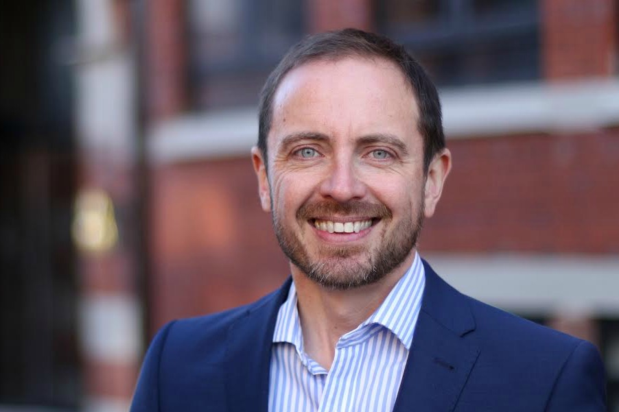 A man in a navy suit and striped shirt is smiling against a background of a brick building.