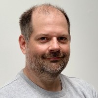 A smiling man with a beard and short hair, wearing a gray t-shirt, against a plain background.