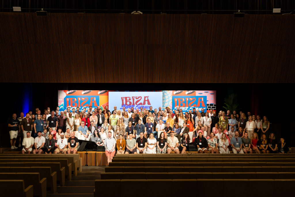 A large group of people poses on a stage in front of a backdrop labeled "IBIZA." The setting appears to be an auditorium with wooden interior and seating visible in the foreground.