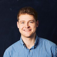 A person with short brown hair smiling, wearing a blue and white striped shirt, with a dark blue background.