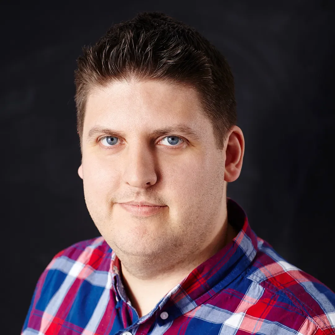 A person with short dark hair wearing a red and blue plaid shirt, looking at the camera with a neutral expression, set against a dark background.