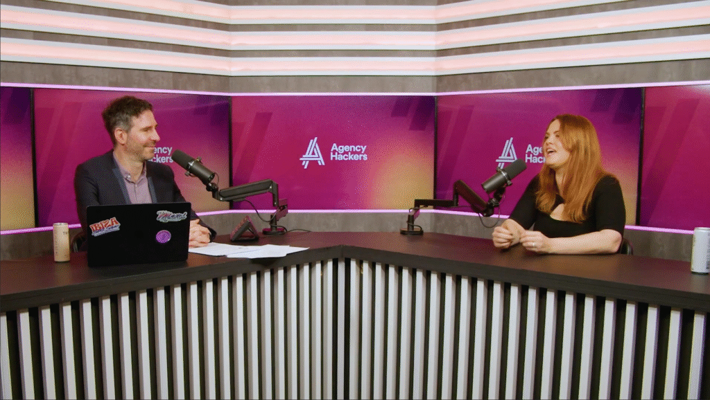 Two people are seated at a podcast table with microphones, engaged in conversation. The studio has a vibrant pink and purple backdrop with podcast branding.