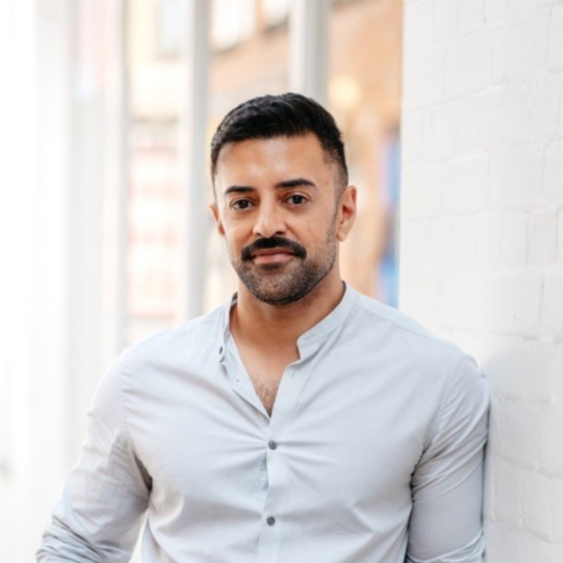 Person with short dark hair and mustache wearing a light button-up shirt, leaning against a white brick wall.