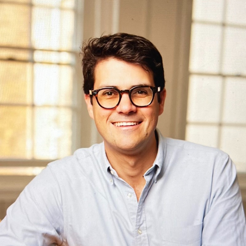 A smiling person with glasses and short dark hair, wearing a light blue shirt, is seated in front of large windows with white frames.