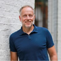 A man in a navy blue polo shirt stands smiling against a white brick wall.
