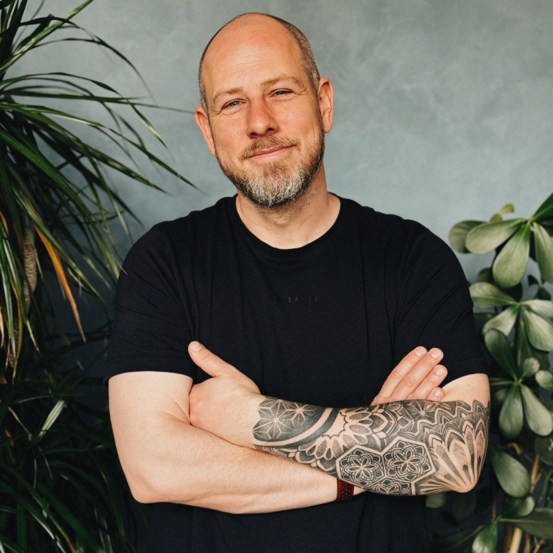 A bearded man with a tattooed arm crosses his arms, standing in front of a plant-filled background.