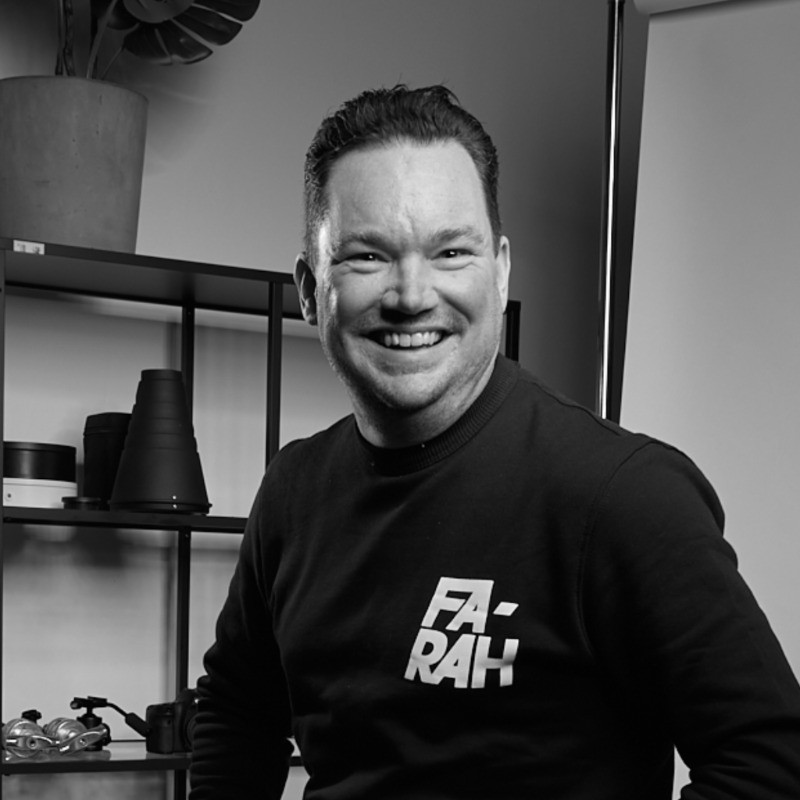 Smiling person in a black sweater stands in a modern room with shelves and decor in the background.