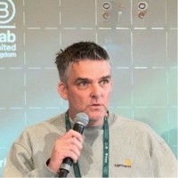 A person wearing a light-colored sweater and lanyard, speaking into a microphone against a backdrop with logos.