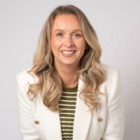 A person with long blonde hair, wearing a white blazer over a green striped shirt, smiles against a plain background.