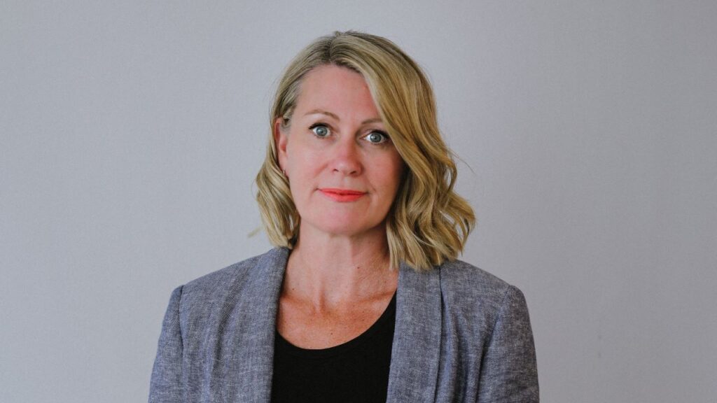 A woman with blonde hair wearing a grey blazer and black top, standing against a neutral background.