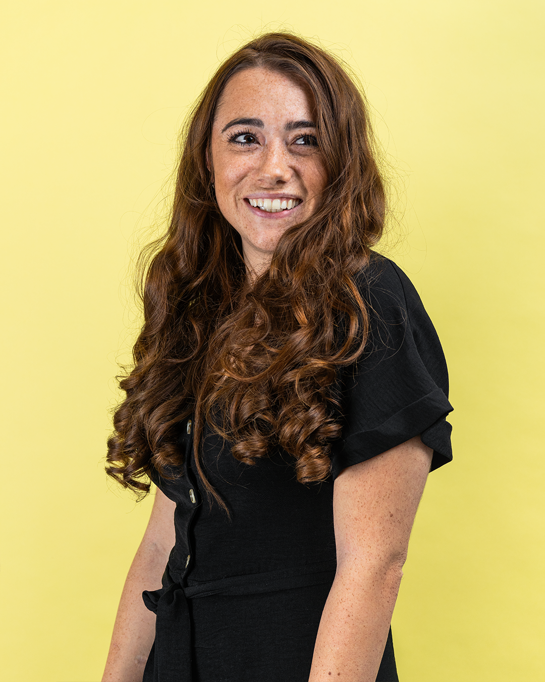Woman with long curly hair smiling in a black dress against a yellow background.
