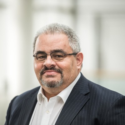 Man with glasses and a graying beard wearing a suit and white shirt, smiling slightly. The background is softly blurred and appears to be an indoor setting.