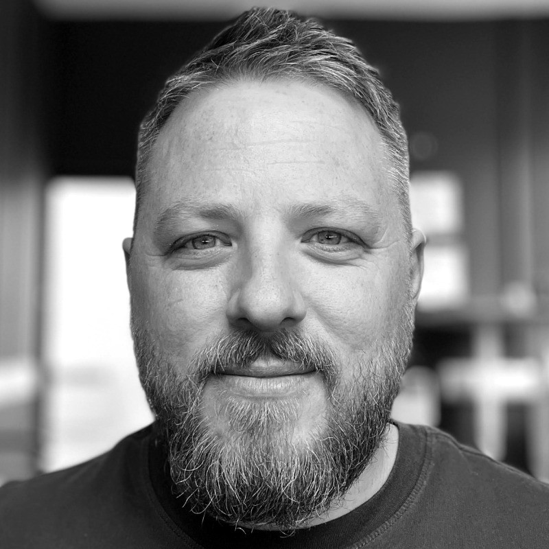 A man with short hair and a beard smiles slightly, captured in black and white.