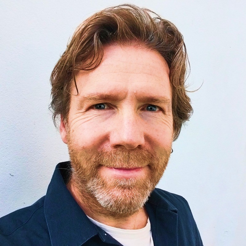 A man with wavy hair and a beard smiles at the camera, wearing a dark shirt against a plain background.