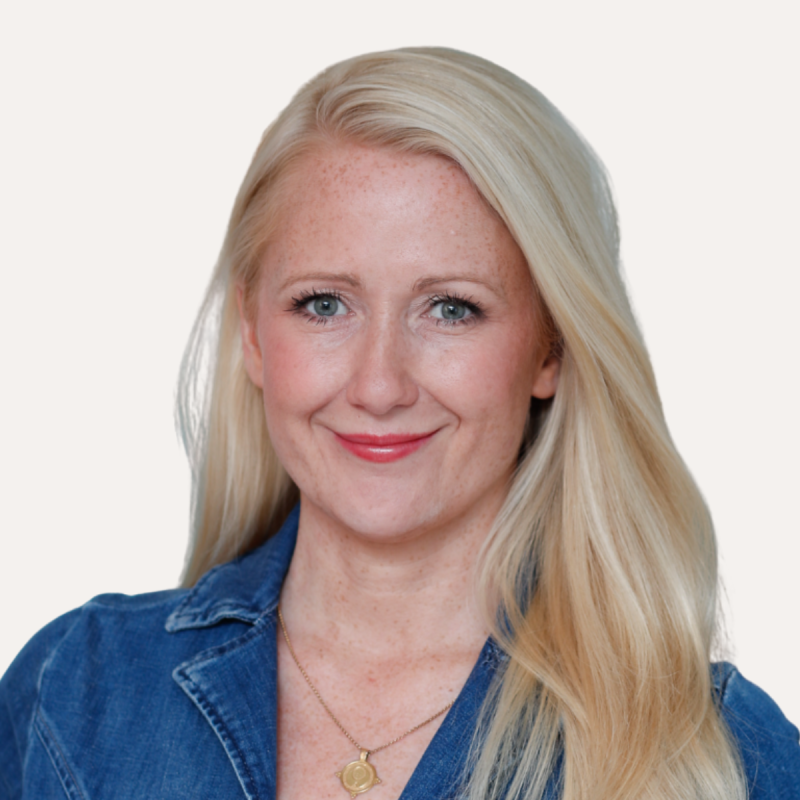 Smiling woman with long blonde hair wearing a denim shirt and a necklace against a plain white background.