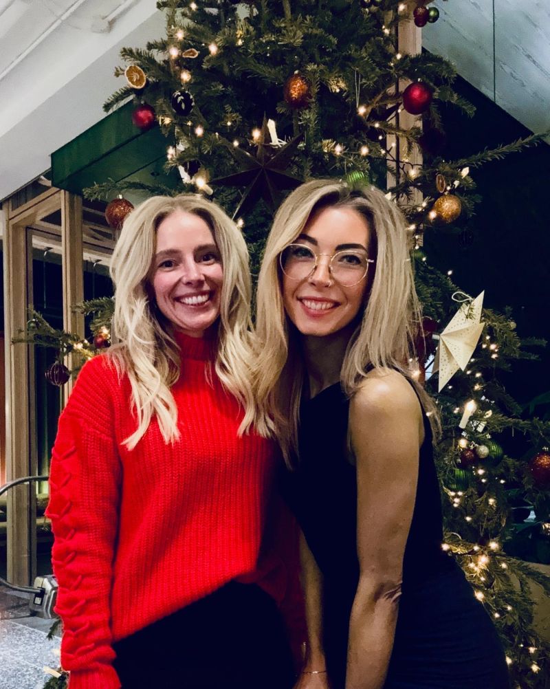 Two women smiling in front of a decorated Christmas tree, one wearing a red sweater and the other in a black outfit.