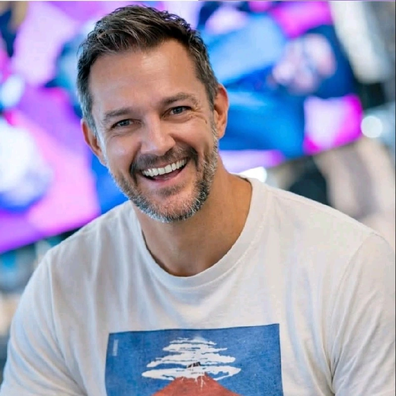 A smiling man with a beard wearing a white T-shirt featuring a colorful mountain design. The background is blurred with vibrant colors.