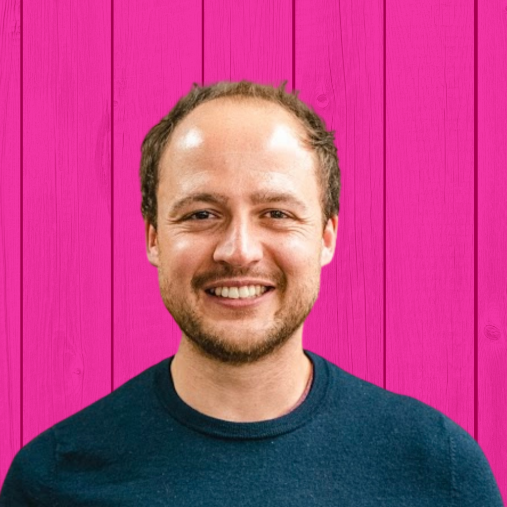 A smiling person with short hair wearing a dark shirt stands against a bright pink wooden background.