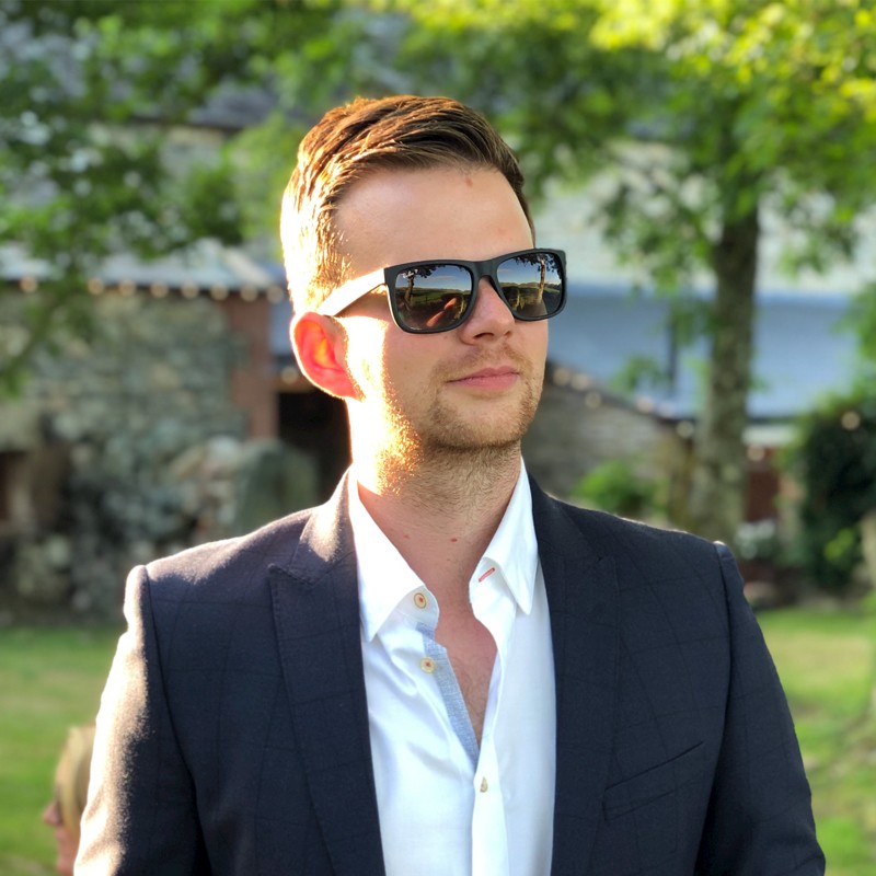 A man in a suit and sunglasses stands outdoors, with trees and a stone building in the background, basking in the sunlight.