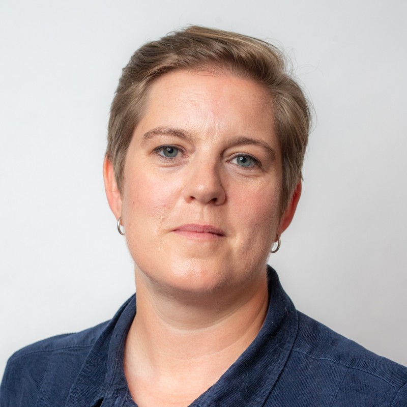 A person with short hair wearing a blue shirt against a plain background, gazing directly at the camera.