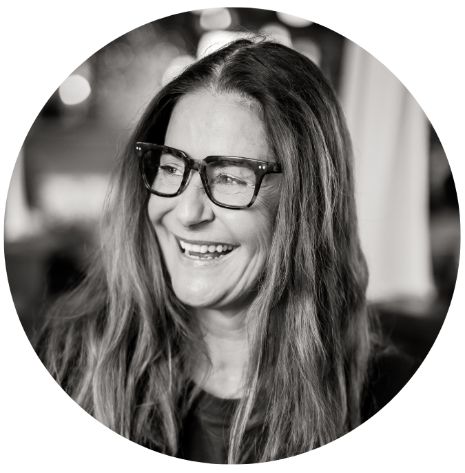 A black and white portrait of a smiling woman with long hair wearing glasses, looking slightly to the side.