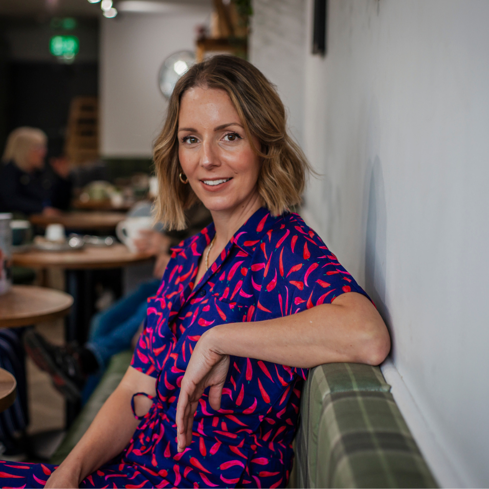 A woman in a patterned dress sitting on a bench in a cafe, smiling at the camera.